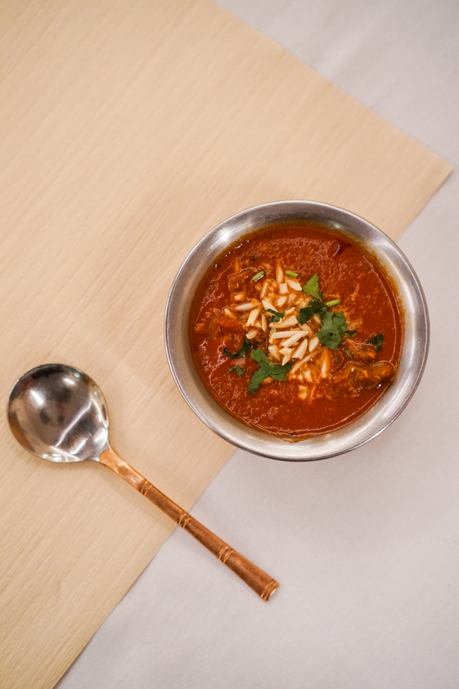 Rogan Josh in a steel bowl, garnished with slivered almonds and fresh coriander on a wooden board.