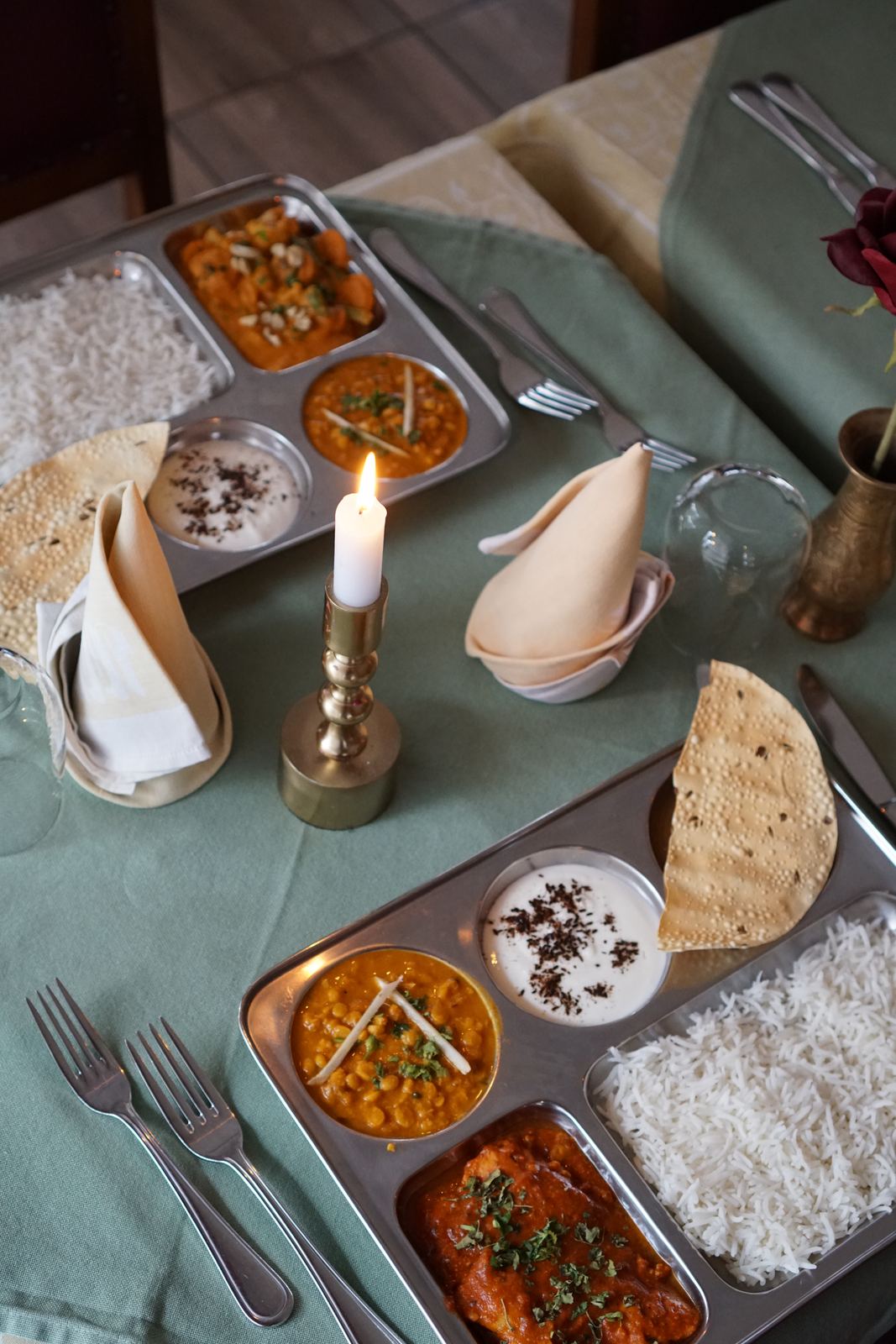 Two thali trays with curry, dal, raita, basmati rice and papad on a set table with a candle.